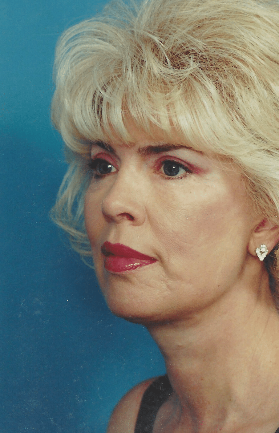 Profile view of a woman with blonde hair styled in soft waves, wearing earrings and subtle makeup, against a blue background.